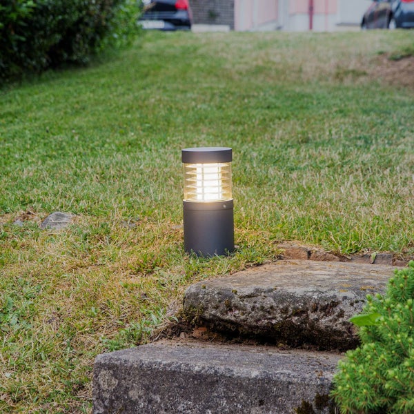 Zylindrische Wegeleuchte aus anthrazitfarbenem Metall mit Lamelleneinsatz am Lichtauslass auf einer Wiese neben Natursteinstufen.