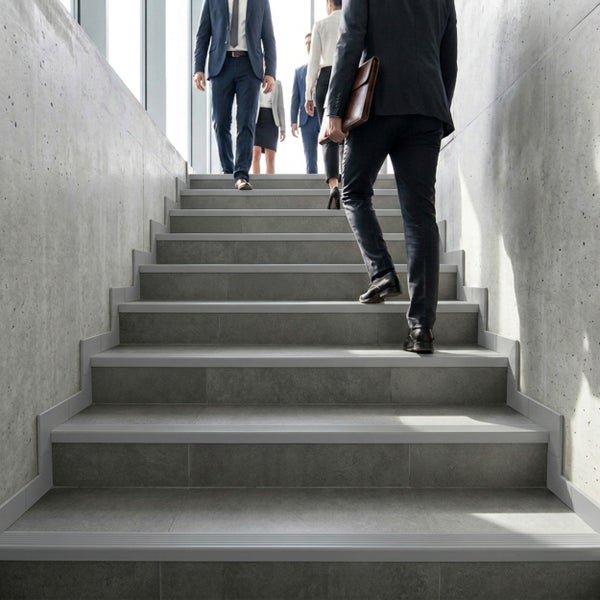 Moderne Treppe mit grauen Fliesen in Natursteinoptik, Treppenkantenprofilen und passenden Sockelleisten an Sichtbetonwänden in einem hellen Bürogebäude.