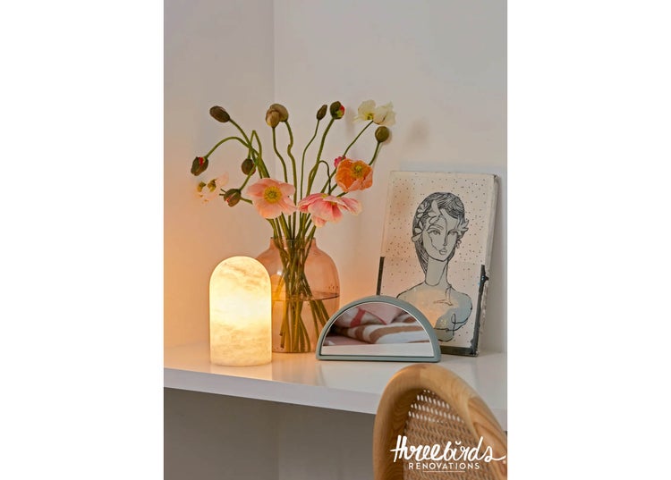 Eine dekorative Tischleuchte neben einer Glasvase mit Mohnblumen, einem halbrunden Tischspiegel und einem gerahmten Kunstbild auf einem Tisch.