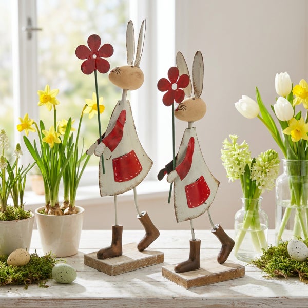 Zwei Osterhasen Figuren aus Metall auf Holzsockel als Dekoration mit Blumen und Zwiebelblumen auf einem Holztisch.