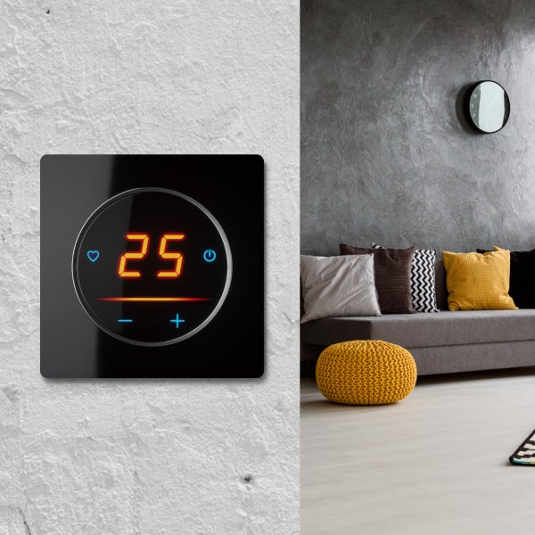 Digitales Raumthermostat mit Touchscreen und Anzeige der Temperatur an einer Wand neben einem Wohnraum mit Sofa und Dekokissen.