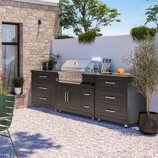 Moderne Außenküche mit schwarzer Schrankkombination, integriertem Edelstahl-Gasgrill und Steinwänden auf hellem Kiesboden.