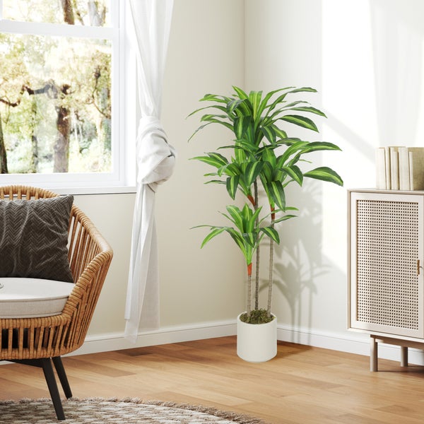 Künstliche Dracaena-Pflanze in weißem Topf, steht in einem hellen Wohnzimmer mit Rattansessel und Holzschrank.