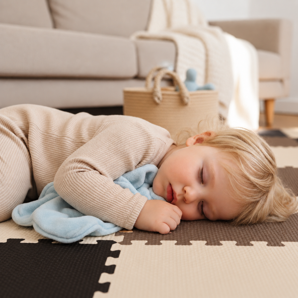 Kleinkind schläft auf einer Puzzlematte aus Schaumstoff im Wohnzimmer vor einem Sofa.