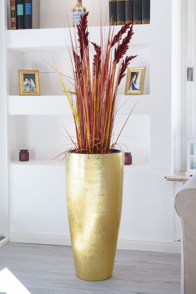 Hohe goldfarbene Bodenvase mit dekorativem Ziergras in einem hell eingerichteten Wohnzimmer vor einem weißen Regal.