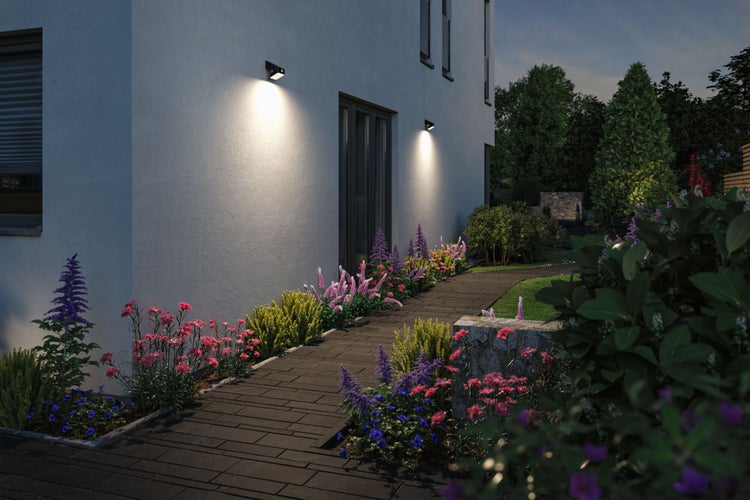 Moderne Außenwandleuchten beleuchten eine Hausfassade und einen gepflasterten Gartenweg mit blühenden Pflanzen in einem gepflegten Garten.