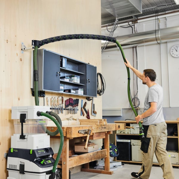 Ein Mann nutzt in einer hellen Werkstatt einen Festool Absaugarm mit Absaugmobil und Saugschlauch über einer Werkbank aus Holz.