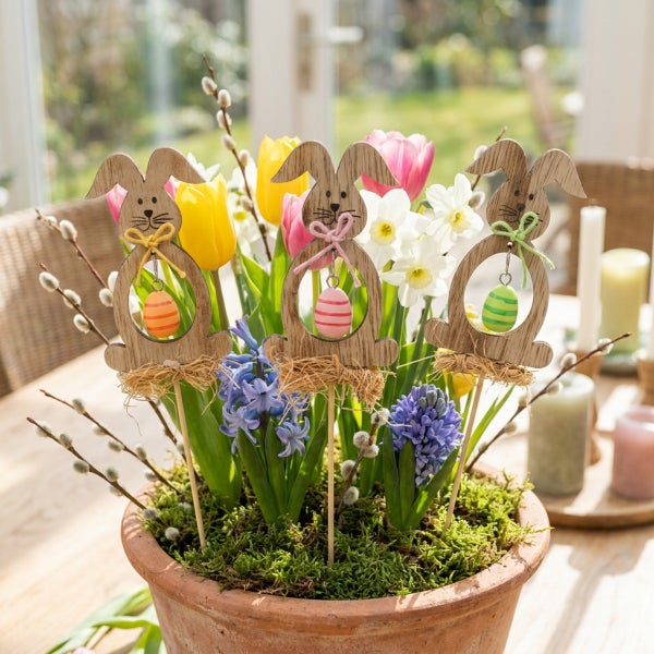Ostergesteck mit drei hölzernen Osterhasen-Steckern, verziert mit kleinen Eiern, in einem Tontopf mit Frühlingsblumen wie Tulpen und Hyazinthen.