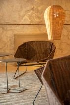 Wohnbereich mit Lounge Sesseln aus Flechtwerk, filigranen Beistelltischen und einer Rattan Hängeleuchte vor einer strukturierten Wand.
