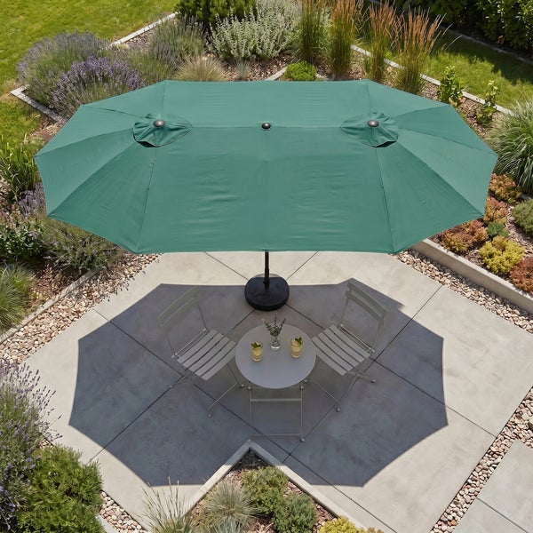 Großer grüner ovaler Gartenschirm über einem runden Tisch und zwei Klappstühlen auf einer Terrasse in einem Garten.