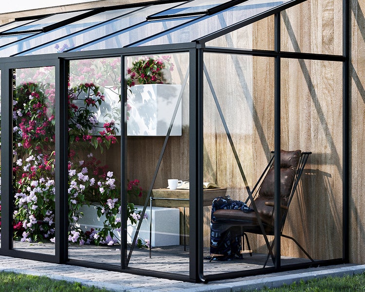 Anlehn-Gewächshaus aus schwarzem Aluminium und Glas an einer Holzwand, eingerichtet mit Pflanzen, Sessel und Beistelltisch auf Steinplatten.