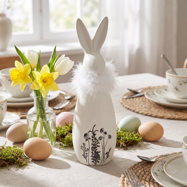 Osterdekoration bestehend aus einer Keramik Osterhasenfigur mit Federkragen und Blumenmuster auf einem gedeckten Ostertisch mit Frühlingsblumen