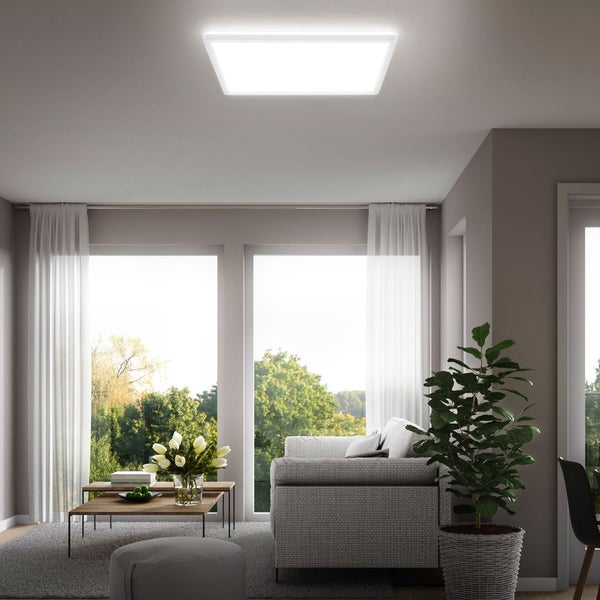 Modernes, helles Wohnzimmer mit quadratischem LED-Panel an der Decke, grauem Sofa, weißen Vorhängen und Blick ins Grüne.