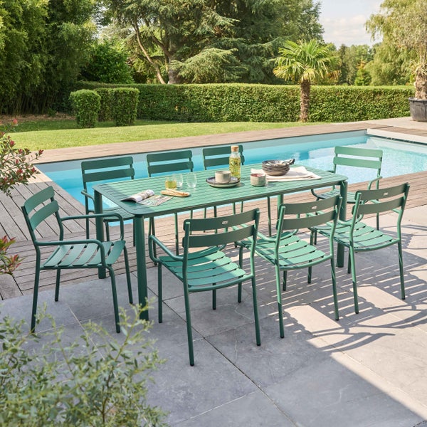 Grünes Gartenmöbel-Set aus Metall mit rechteckigem Tisch und acht Stühlen auf einer hellen Steinterrasse an einem Pool in einem Garten.