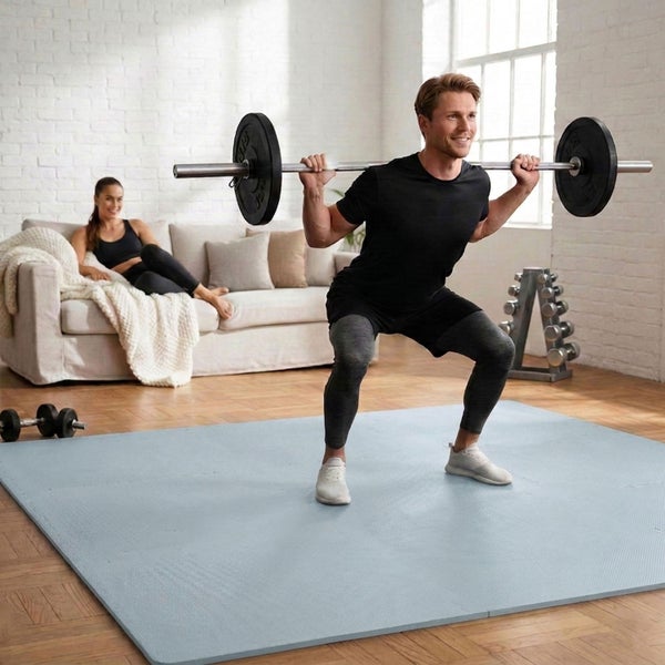 Mann macht Kniebeugen mit einer Langhantel auf einer Bodenschutzmatte in einem hellen Wohnzimmer mit Fitnessgeräten.