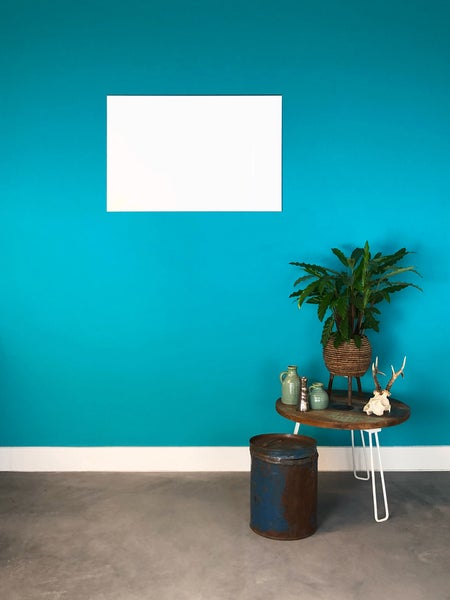 Innenraum mit einer türkis gestrichenen Wand, weißer Sockelleiste und einem runden Holztisch mit Zimmerpflanze neben einem Metallbehälter.