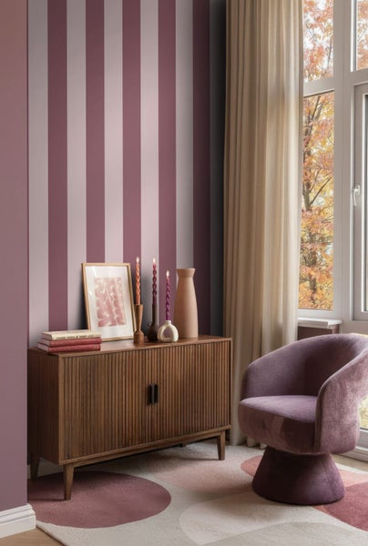 Vertikal gestreifte Tapete in Rosa- und Mauvetönen hinter einem Sideboard aus Holz und einem Sessel in einem hellen Wohnraum.