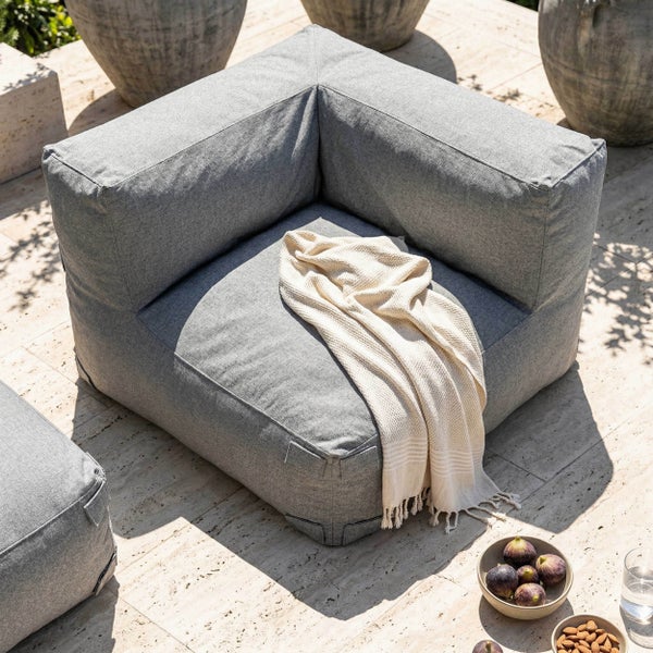 Quadratisches Eckelement für eine Outdoor-Lounge aus grauem Stoff mit einer hellen Decke auf einer Steinterrasse.