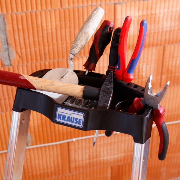 Werkzeug auf einer Trittleiter mit Krause Logo, darunter Hammer, Maurerkelle und Zangen vor einer Ziegelwand
