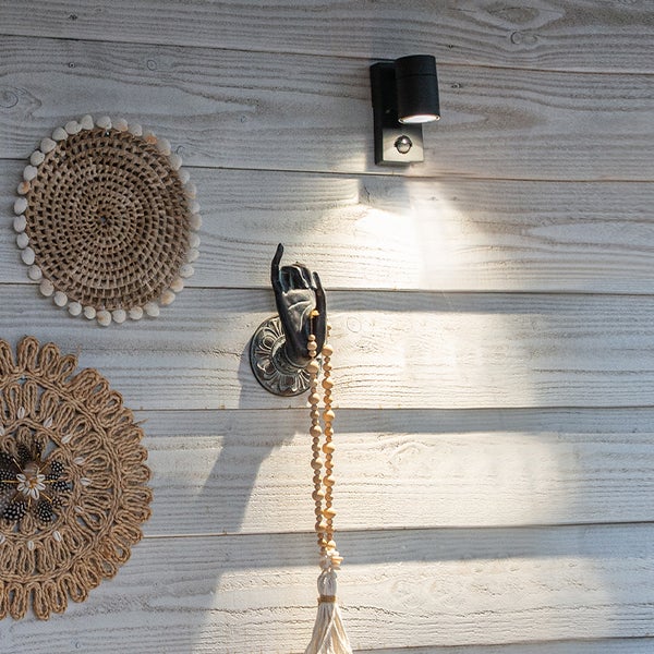 Schwarze Außenwandleuchte mit Bewegungsmelder an einer hellen Holzwand neben runder Wanddekoration und einem Wandhaken in Handform.