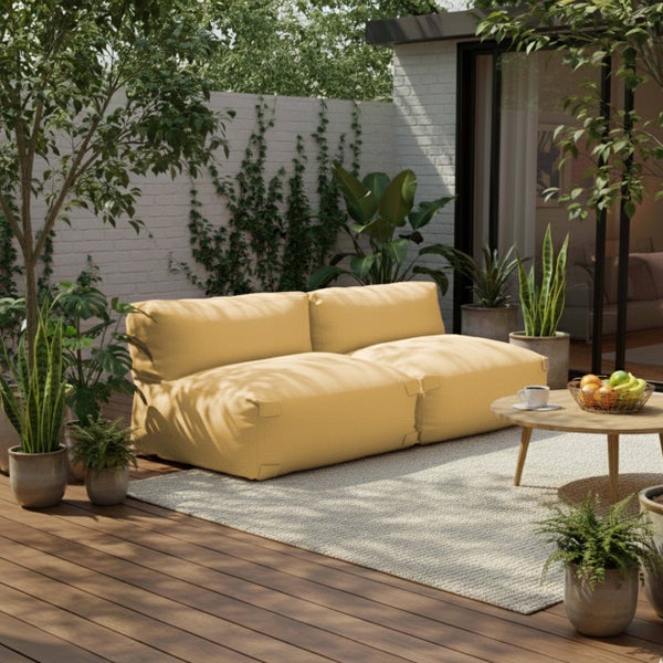 Modernes Outdoor-Loungesofa in Senfgelb auf einer Holzterrasse mit rundem Holztisch, hellem Teppich und vielen Grünpflanzen. Helle Atmosphäre.