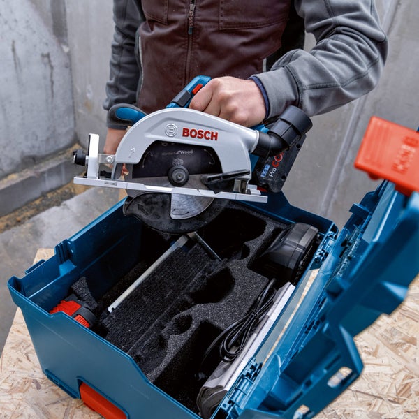 Eine Person entnimmt eine Bosch Akku-Handkreissäge aus einem blauen Werkzeugkoffer auf einer Baustelle.