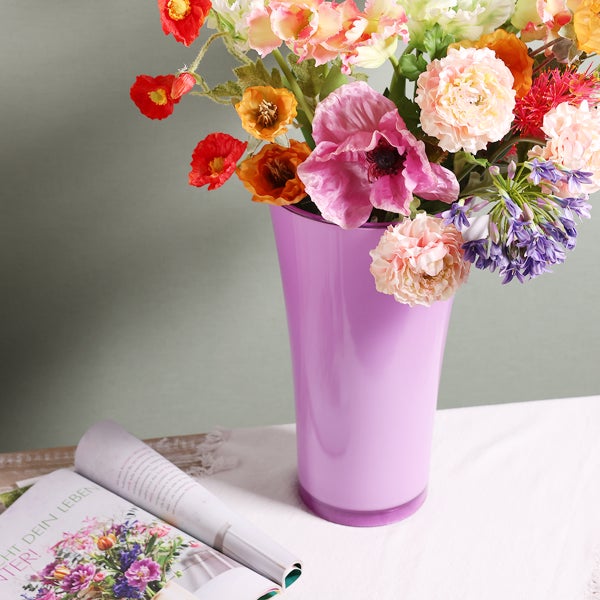 Lilafarbene Glasvase mit einem bunten Strauß Kunstblumen auf einem weißen Tisch.