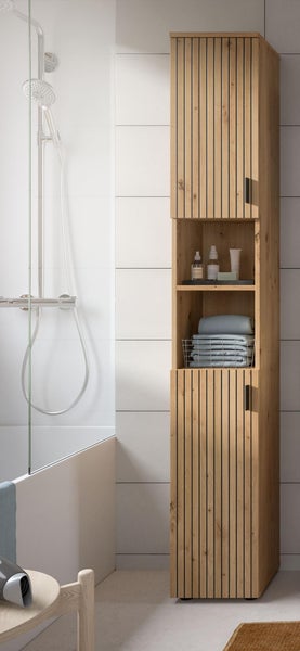 Schmaler Badhochschrank in Eichenoptik mit Lamellendesign, zwei Türen und offenen Fächern, stehend in einem hellen Bad neben einer Dusche.