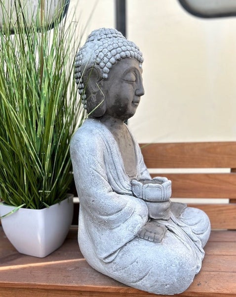 Sitzende Buddha-Figur für den Garten, eine Schale haltend, in Steinoptik, seitlich auf einer Bank neben einer Pflanze.