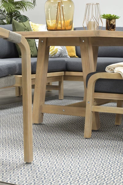 Gartentisch aus Holz mit grauer Lounge-Sitzgruppe, Zierkissen und gemustertem Outdoor-Teppich.