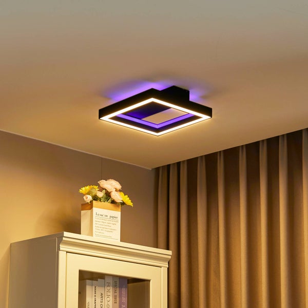 Moderne quadratische LED-Deckenleuchte mit warmweißem Licht und lila Hintergrundbeleuchtung über einem hellen Schrank mit Blumen und Vorhängen.