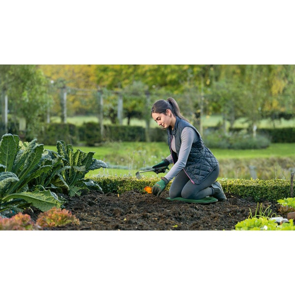 Eine Frau kniet auf einem Kniekissen und arbeitet mit einer Harke im Garten.