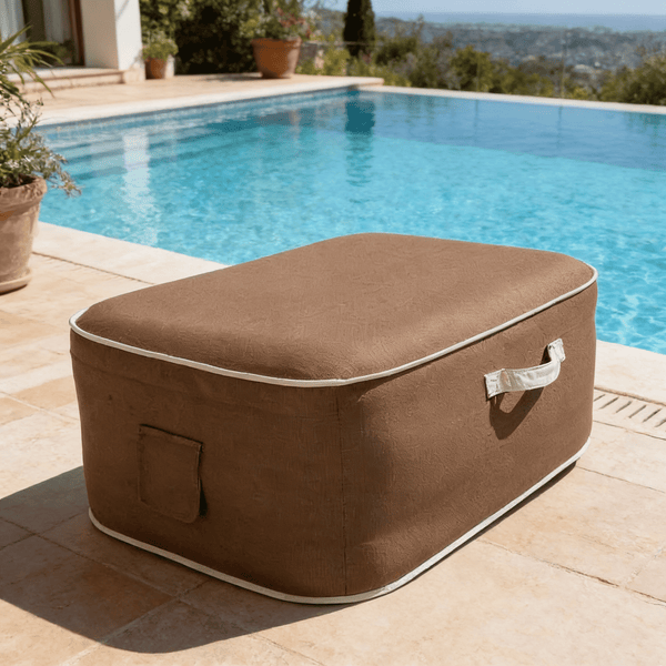 Rechteckiger brauner Outdoor Sitzsack mit weißer Paspelierung und Tragegriff auf einer Terrasse neben einem Pool.