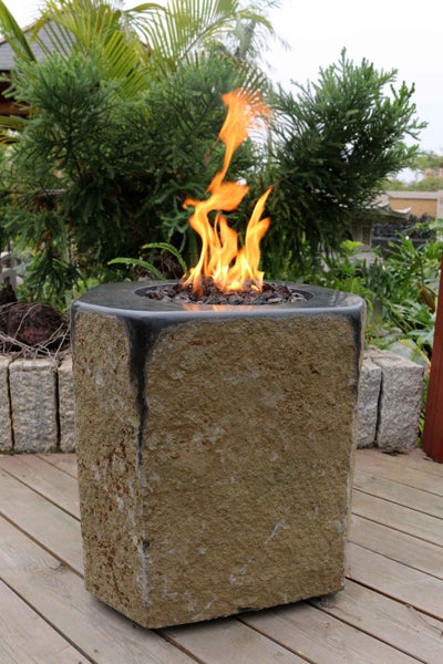 Gaskamin für den Außenbereich aus Naturstein mit Flammen auf einer Holzterrasse inmitten von Pflanzen.