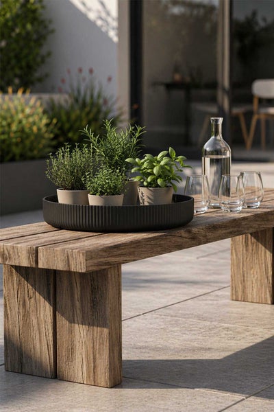Rundes, geriffeltes Tablett mit Kräutertöpfen auf einer rustikalen Holzbank auf einer Terrasse neben einer Glaskaraffe.
