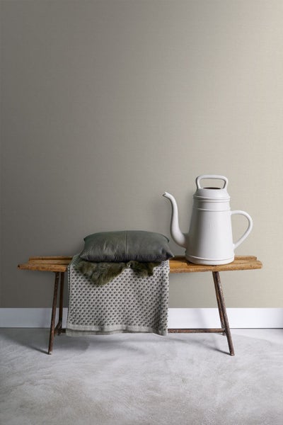 Dekorative Szene mit Bank, Kissen, Decke und Gießkanne vor einer strukturierten Wand