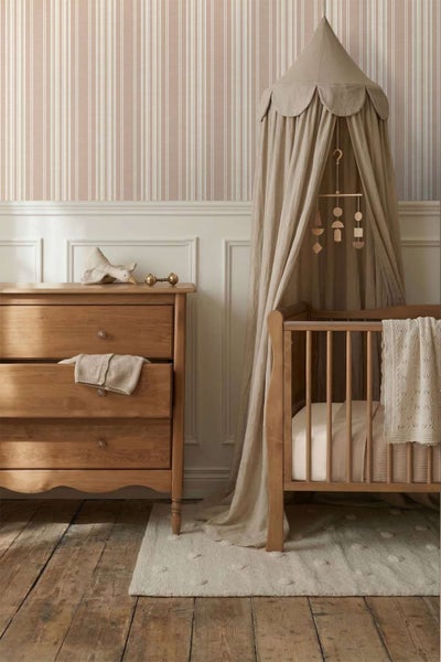 Ein Kinderzimmer mit gestreifter Tapete, einem Babybett mit Betthimmel, einer Kommode aus Holz und einem Teppich auf einem Holzdielenboden.