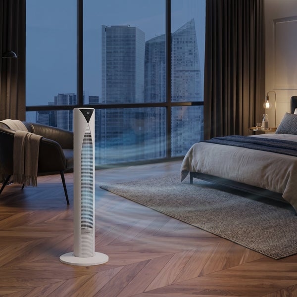 Weißer Turmventilator in einem Schlafzimmer mit Parkettboden, Bett und großem Fenster mit Blick auf die nächtliche Stadt.