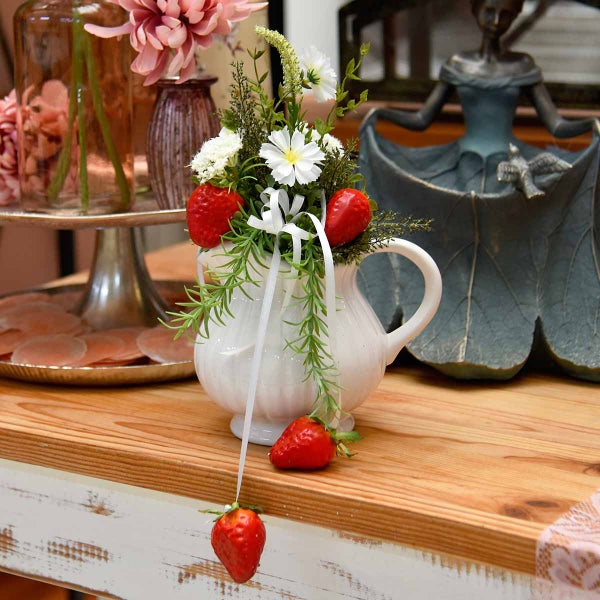 Dekorative Blumen und Erdbeeren in einer weißen Krugvase auf einem Holztisch.