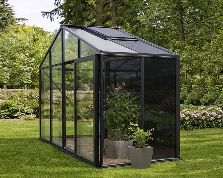 Modernes Gewächshaus mit anthrazitfarbenem Metallrahmen und Glaspaneelen in einem hellen, geordneten Garten mit Rasen, Bäumen und Topfpflanzen.