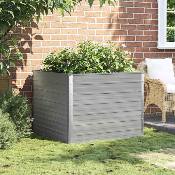 Quadratisches Hochbeet aus Metall in einem Garten vor einer Ziegelwand, bepflanzt mit grünen Pflanzen.