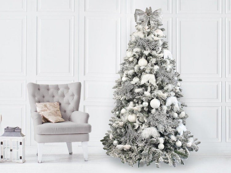 Künstlicher Weihnachtsbaum mit Schneeoptik, dekoriert mit hellen Kugeln und einer großen Schleife, neben einem Sessel in einem hellen Raum.