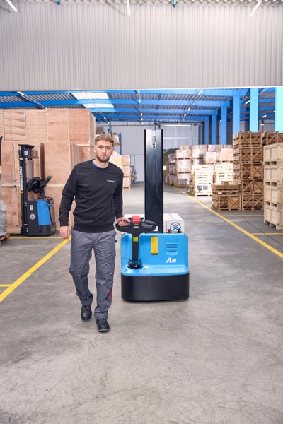 Ein Mitarbeiter in einem Lager bedient einen blauen Elektro-Deichselstapler zwischen Holzkisten.