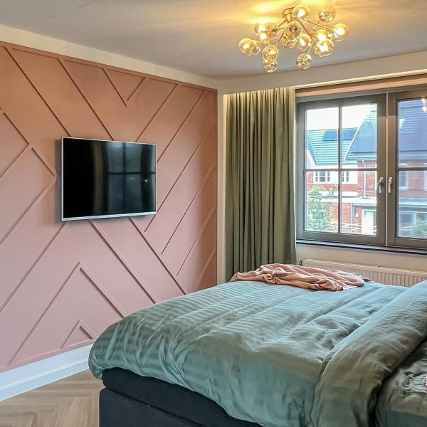Helles Schlafzimmer mit rosa geometrischer Wand, wandmontiertem Fernseher, grüner Bettwäsche, grünen Vorhängen und goldener Deckenleuchte.
