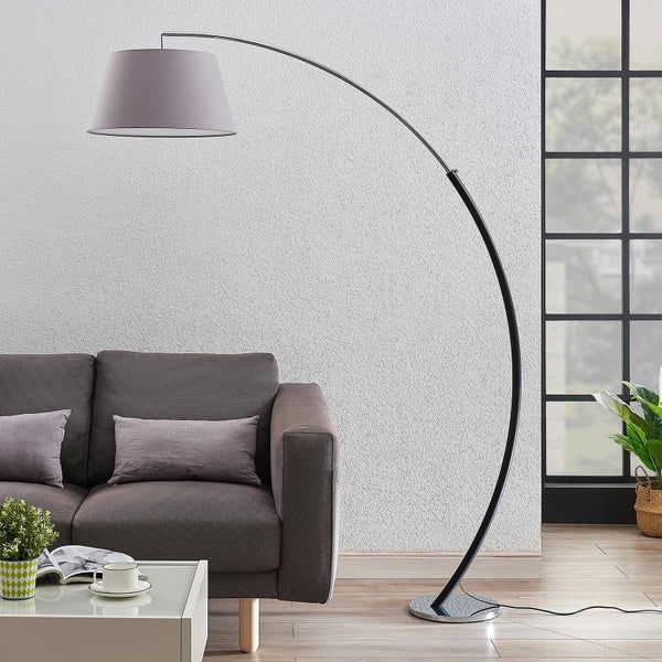 Moderne Bogenlampe mit Stoffschirm und gebogenem Metallgestell in einem hellen Wohnzimmer neben einem Sofa.
