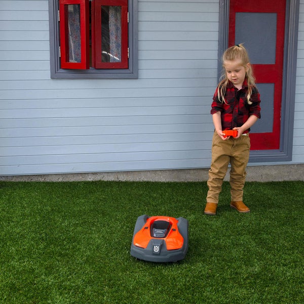 Ein Kind mit einer Fernbedienung steht neben einem Rasenmähroboter auf einer Rasenfläche vor einem Haus.