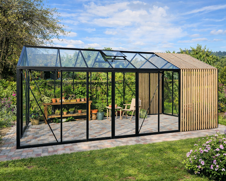Modernes Gewächshaus mit schwarzem Aluminiumrahmen, Glaswänden und Holzlamellen-Anbau auf gepflasterter Fläche im Garten.