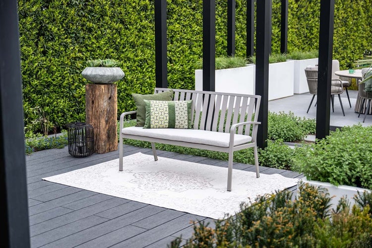 Moderne Gartenbank aus Metall mit Sitzkissen und Lamellen-Rückenlehne auf einer Terrasse mit Outdoor-Teppich und Pflanzen.