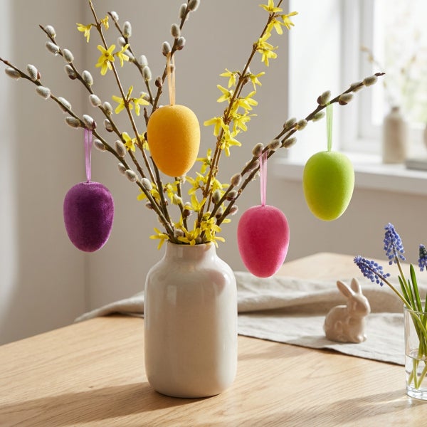 Osterstrauß aus Weidenkätzchenzweigen und Forsythien in einer Vase mit hängenden, bunten Ostereiern aus Textil auf einem Holztisch.