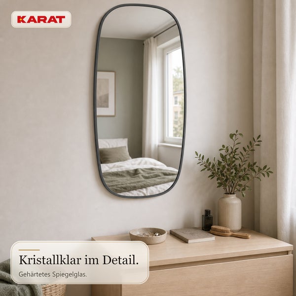 Ovaler Wandspiegel mit schwarzem Rahmen von Karat Logo, montiert über einer hölzernen Kommode in einem hellen Schlafzimmer mit gehärtetem Spiegelglas.